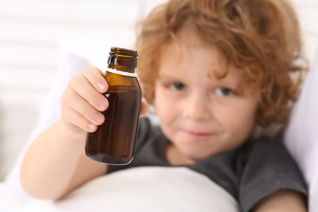 Cute boy holding cough syrup in bed, focus on bottleの写真素材