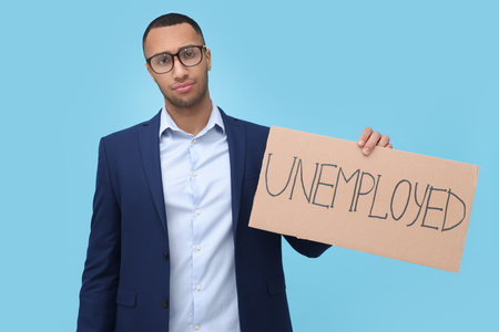 Young man holding sign with the word Unemployed on light blue backgroundの写真素材