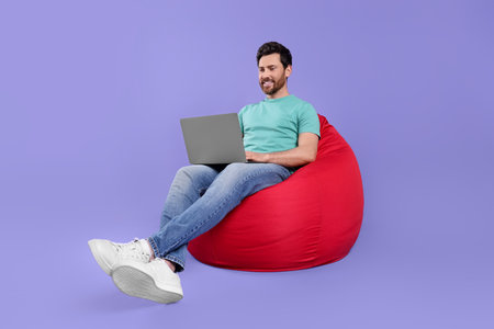 Happy man with laptop sitting on beanbag chair against purple backgroundの写真素材