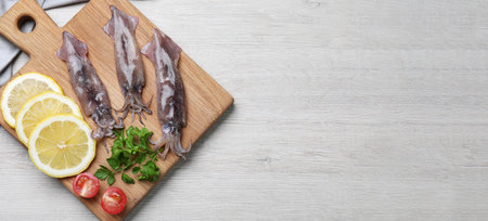 Board with fresh squids, parsley, slices of lemon and tomato on white wooden table, flat lay. Banner...