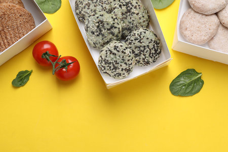 Many different raw vegan meat products, tomatoes and spinach on yellow background, flat lay. Space...