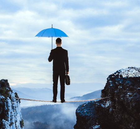 Risks and challenges of entrepreneurship. Businessman with portfolio and umbrella standing on rope over abyss in mountains, back viewの写真素材