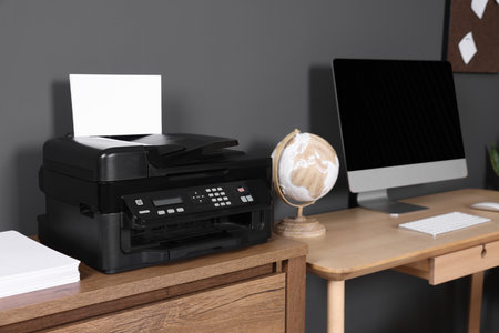 Modern printer with paper near computer on wooden desk at home