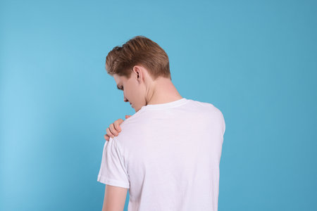 Teenage boy suffering from pain in shoulder on light blue background, back view. Arthritis symptomの写真素材