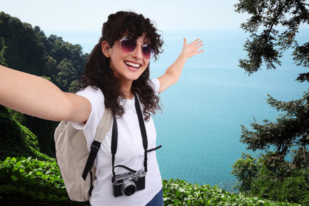 Beautiful woman in sunglasses with camera taking selfie on hill near seaの写真素材