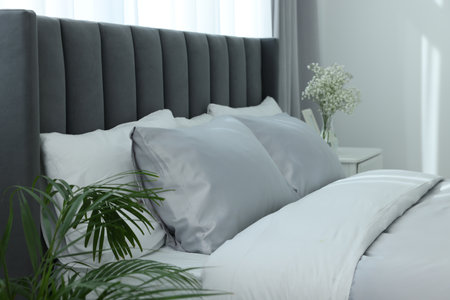 Comfortable bed and vase with beautiful flowers on white bedside table in room. Stylish interior