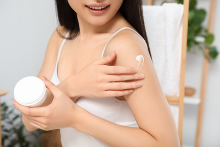 Young woman applying body cream on shoulder in room, closeupの写真素材