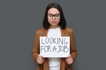 Upset young unemployed woman holding sign with phrase Looking For A Job on grey backgroundの写真素材