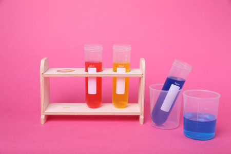 Beakers and test tubes with colorful liquids in wooden stand on bright pink background. Kids...