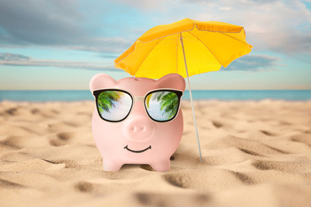 Vacation savings. Piggy bank near umbrella on sandy beach near sea. Reflection of palm leaves in sunglassesの写真素材