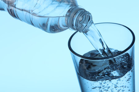 Pouring water from bottle into glass on light blue background, closeup