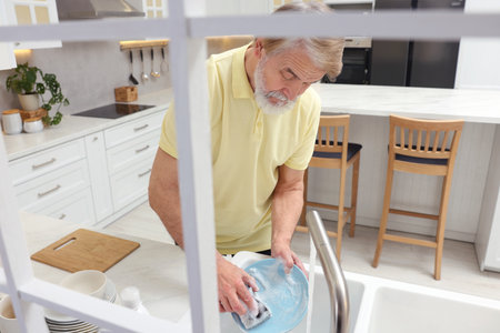 Senior man washing plate above sink in kitchen, view from outsideの写真素材