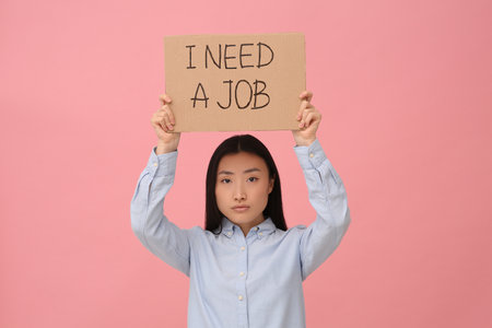 Unemployed Asian woman holding sign with phrase I Need A Job on pink backgroundの写真素材