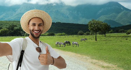 Smiling young man in straw hat taking selfie and showing thumbs up at nature parkの写真素材