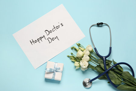 Card with phrase Happy Doctor's Day, stethoscope, gift box and eustoma flowers on light blue background, flat layの写真素材