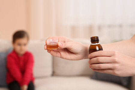 Mother with cough syrup for her daughter in room, focus on handsの写真素材