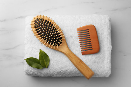 Wooden hair comb, brush, terry towel and green leaves on white marble table, top view