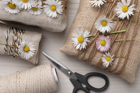 Gifts packed in burlap fabric with beautiful chamomiles, ropes and scissors on white wooden table, flat layの写真素材
