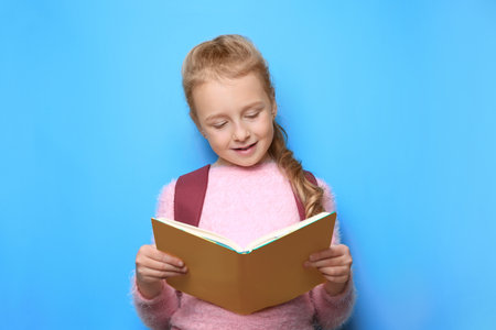 Happy little girl with backpack reading book on light blue background