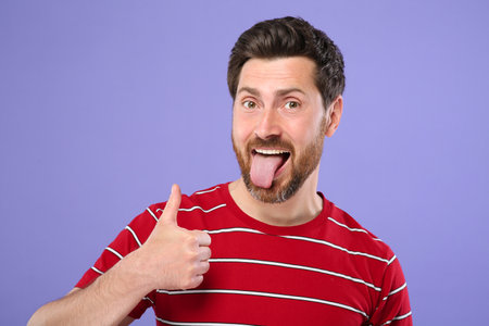 Man showing his tongue and thumbs up on violet background