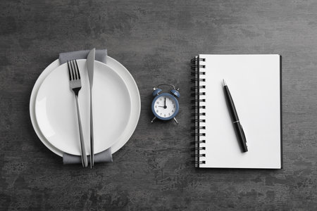 Business lunch concept. Plates, cutlery, alarm clock, notebook and pen on gray textured background,...
