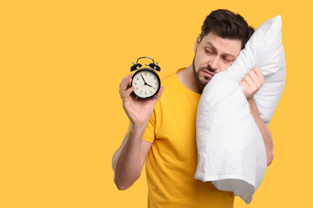 Tired man with alarm clock and pillow on yellow background, space for text. Insomnia problemの写真素材