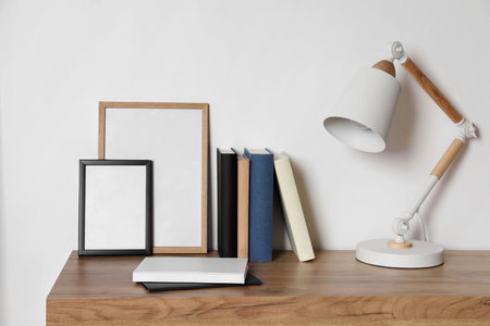 Stylish modern desk lamp, books and frames on wooden table near white wall indoorsの写真素材
