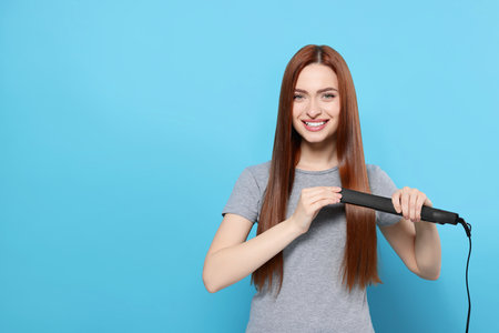 Beautiful woman using hair iron on light blue background, space for textの写真素材