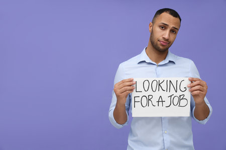 Young unemployed man holding sign with phrase Looking For A Job on violet background. Space for textの写真素材