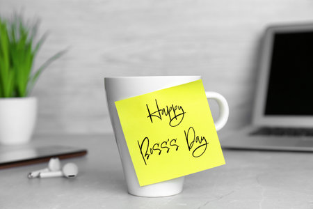 Sticky note with phrase Happy Boss`s Day attached to cup on gray table in officeの写真素材