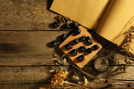 Many black rune stones, old book and dried plants on wooden table, flat lay. Space for text