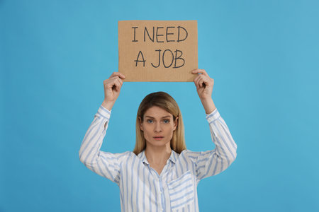 Unemployed woman holding sign with phrase I Need A job on light blue backgroundの写真素材