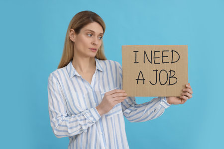 Unemployed woman holding sign with phrase I Need A job on light blue background
