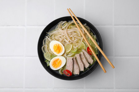 Bowl of delicious rice noodle soup with meat and egg on white tiled table, top viewの写真素材