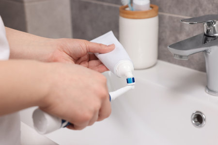 Woman squeezing toothpaste from tube onto toothbrush above sink in bathroom, closeup