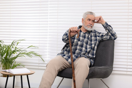 Senior man with walking cane sitting on armchair at homeの写真素材