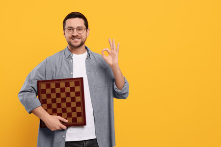Smiling man holding chessboard and showing OK gesture on orange background. Space for textの写真素材
