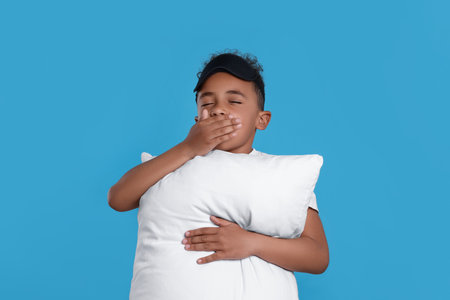 Boy with pillow and sleep mask yawning on light blue background. Insomnia problemの写真素材
