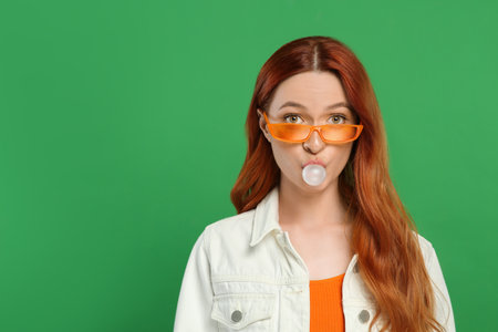Beautiful woman in sunglasses blowing bubble gum on green background. Space for textの写真素材