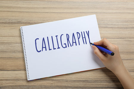 Woman writing word Calligraphy in notebook at wooden table, top view
