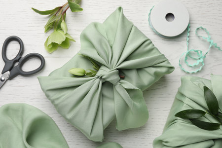 Furoshiki technique. Gifts packed in green fabrics, hellebore flowers, scissors and decorative ribbon on white wooden table, flat layの写真素材