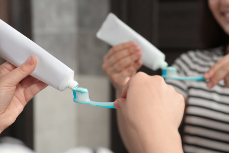 Woman squeezing toothpaste from tube onto toothbrush near mirror in bathroom, closeup