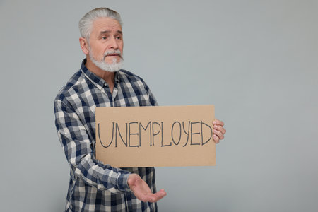 Confused senior man holding cardboard sign with the word Unemployed on light gray background. Space for textの写真素材