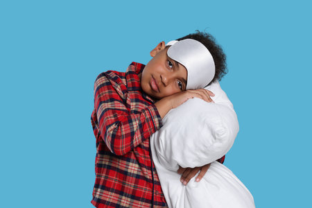 Boy with pillow and sleep mask on light blue background. Insomnia problemの写真素材