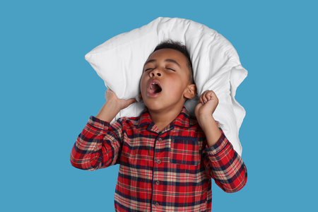 Boy covering his ears with pillow and yawning on light blue background. Insomnia problemの写真素材