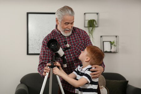 Little boy with his grandfather using telescope in room at homeの写真素材
