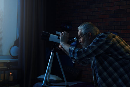 Senior man looking at stars through telescope in roomの写真素材