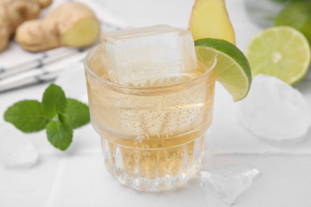 Glass of tasty ginger ale with ice cube and ingredients on white table, closeup
