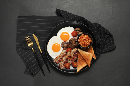 Plate with fried eggs, mushrooms, beans, bacon, tomatoes and toasted bread on black table, flat lay. Traditional English breakfastの写真素材