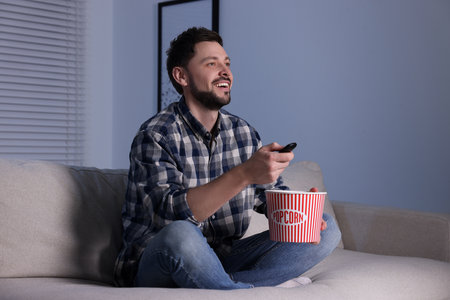Happy man with popcorn bucket changing TV channels with remote control at home in eveningの写真素材